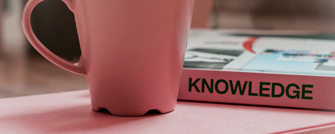 Pink cup on the pink table, behind there is a pink book titled Knowledge